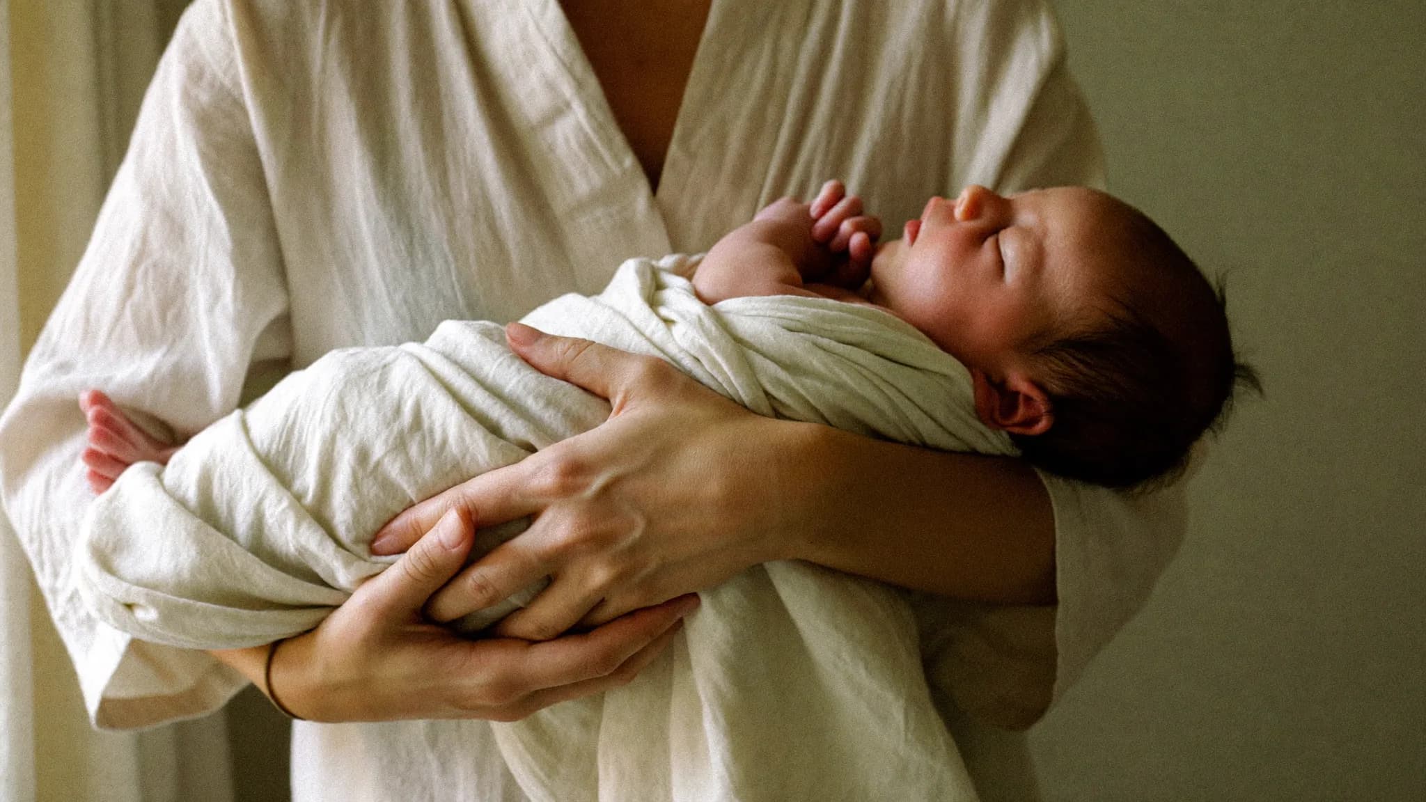 Yeni doğmuş bir bebek, krem renkli muslin bir örtüye sarılmış, annesinin kollarında sakin bir pencere ışığı altında dinleniyor